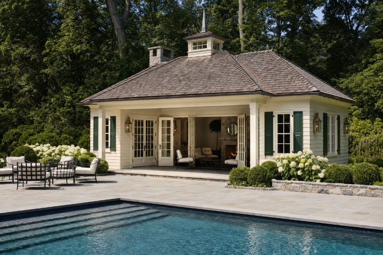 Pool house with white clapboard, green shutters, cedar shingle roof with cupola, open French doors to lounge with stone fireplace, bluestone terrace, pool steps, and white hydrangea borders at the Hydrangea Estate in Greenwich