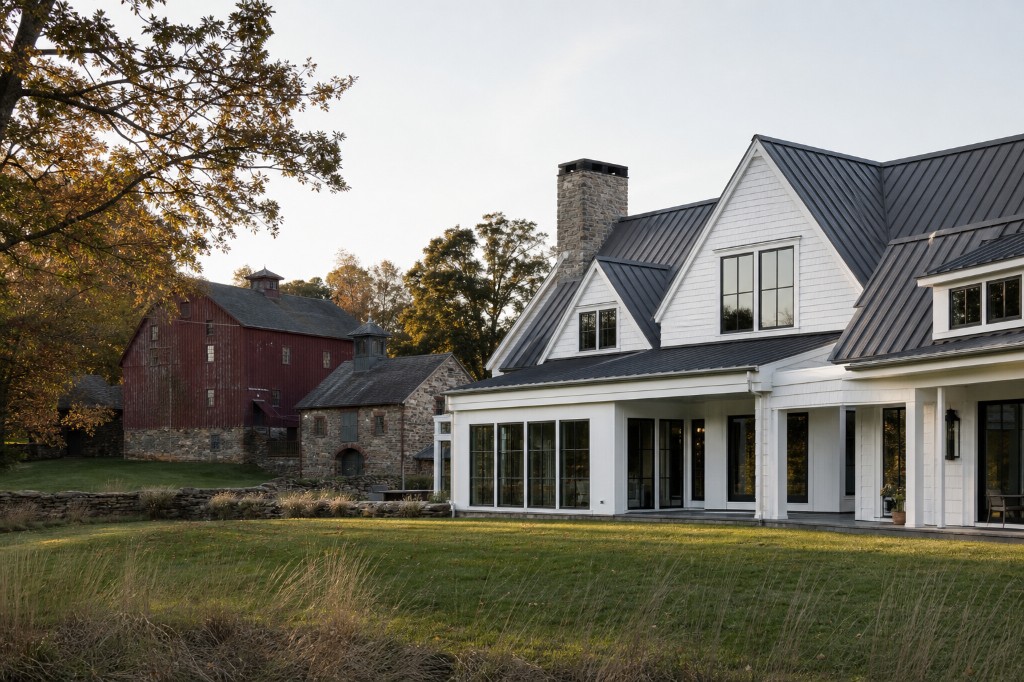 Modern farmhouse with white shingle and clapboard siding, charcoal standing-seam metal roof, fieldstone chimney, and historic red barn and stone outbuildings on an estate lawn in Greenwich